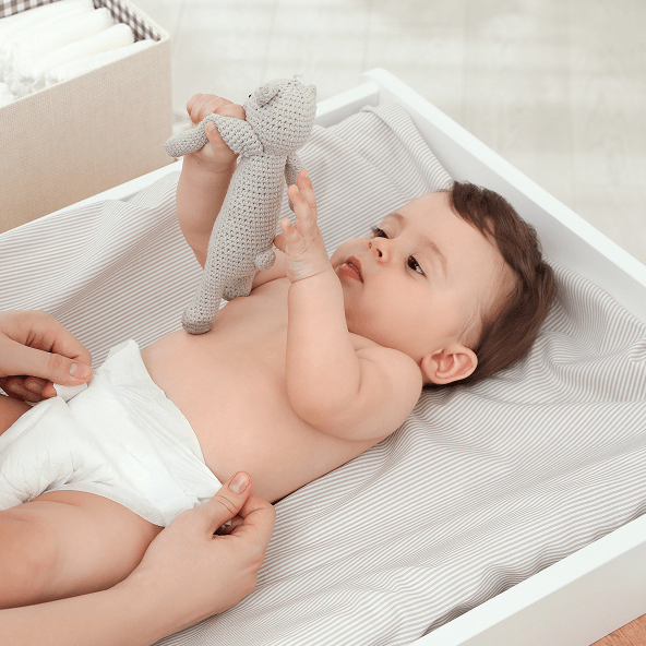 Małe dziecko leży na przewijaku, trzyma w uniesionych rękach zabawkę.