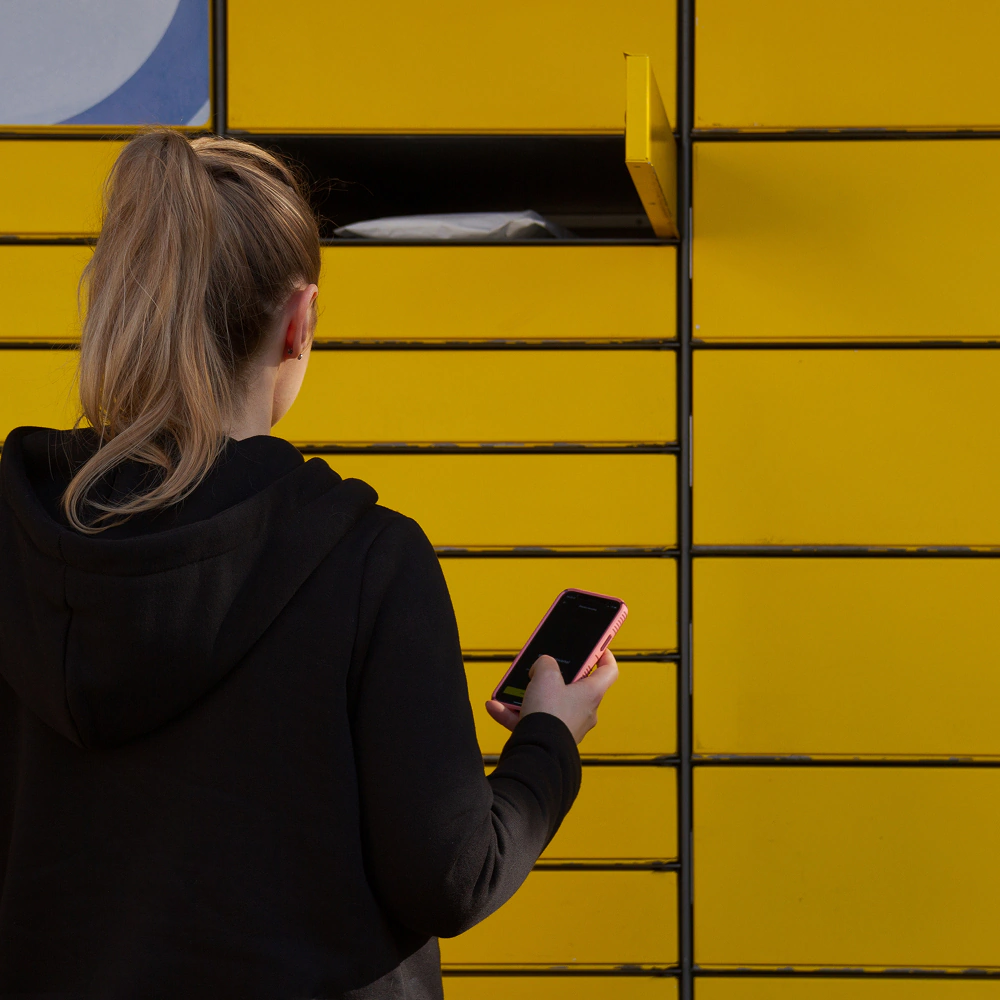 Kobieta z blond włosami upiętymi w kucyk stoi tyłem, w prawym ręku trzyma telefon i odbiera paczkę z otwartego okienka w żółtym paczkomacie.
